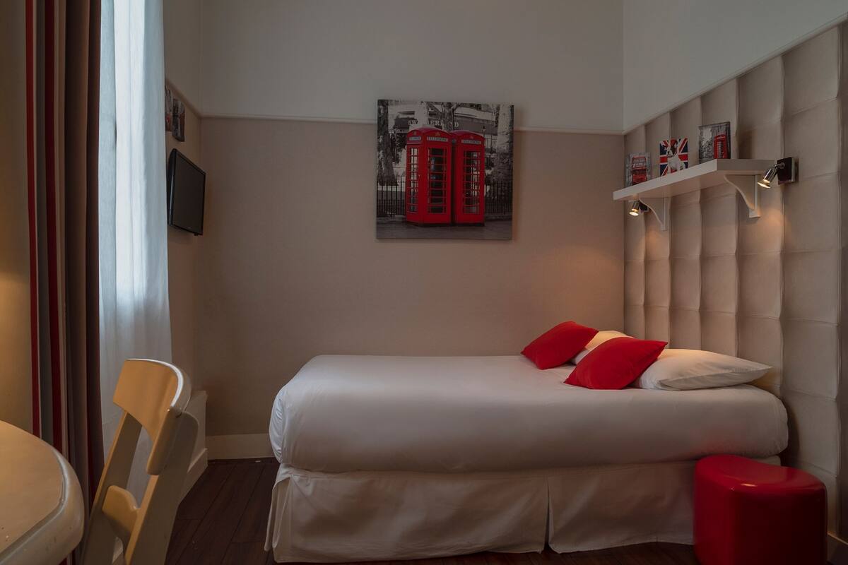 Une chambre d’hôtel avec un lit, une œuvre d’art représentant une cabine téléphonique rouge, une étagère avec des livres et une chaise.
