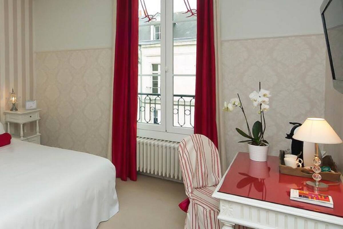 Une chambre d’hôtel avec un lit, une table de chevet, une chaise, une fenêtre avec des rideaux rouges et une vue sur les bâtiments environnants.