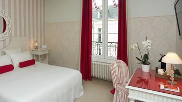 Ett hotellrum med en säng, ett nattduksbord, en stol, ett fönster med röda gardiner och utsikt över byggnader utanför.