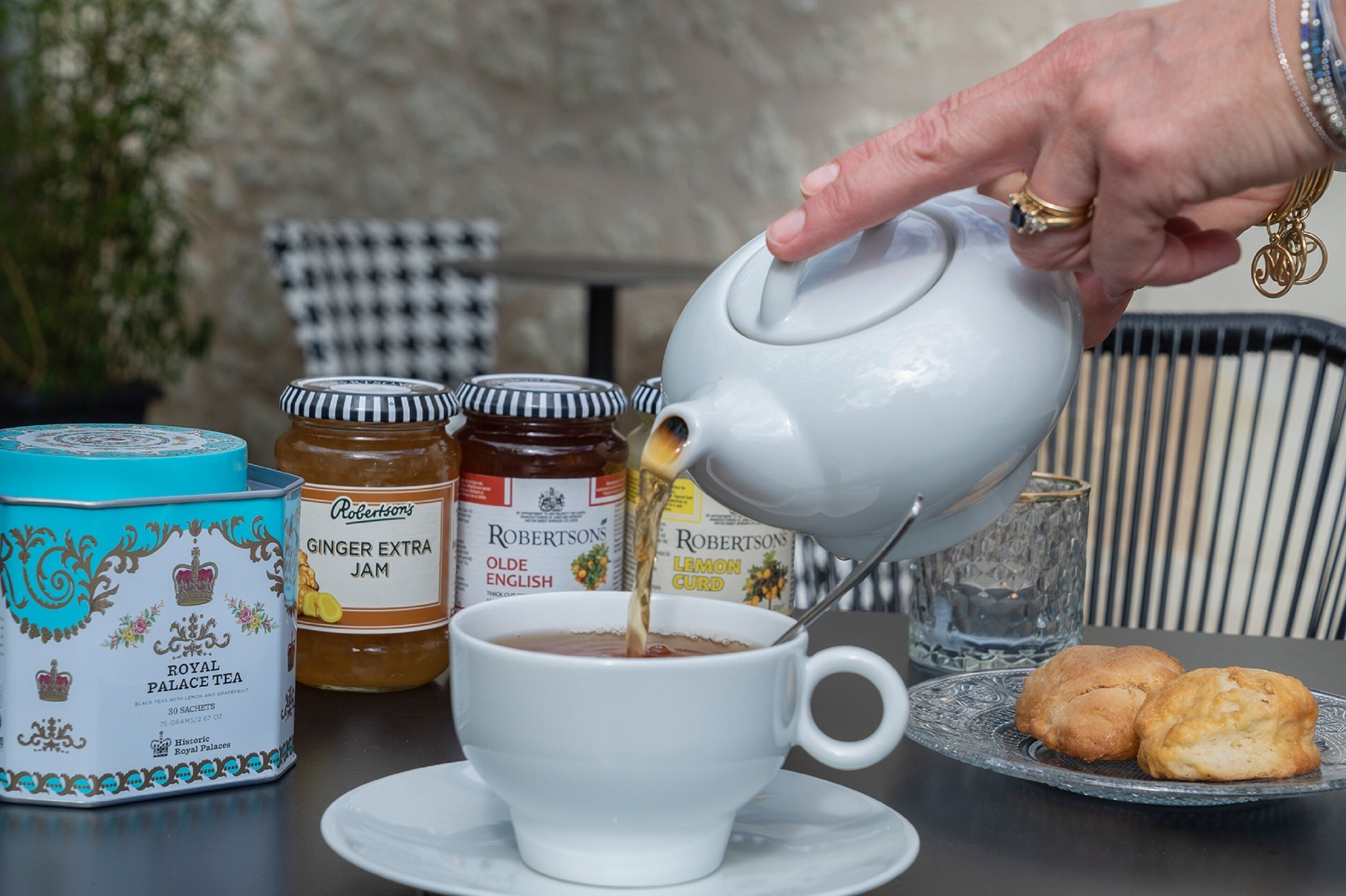 Una mano vertiendo té de una tetera a una taza, con frascos de mermelada y una caja de té con la etiqueta 'Royal Palace Tea' sobre la mesa.