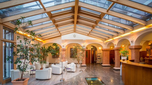 A spacious lobby with a high ceiling, wooden beams, and a glass roof. There are several seating areas with white sofas and potted plants.