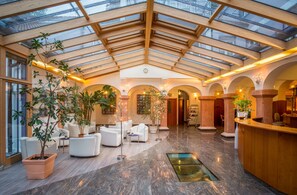 Un hall d’entrée spacieux avec un plafond haut, des poutres en bois et un toit en verre. On y trouve plusieurs coins salon avec des canapés blancs et des plantes en pot.