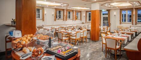 Uma sala de jantar de hotel com várias mesas e cadeiras, um buffet com diversos pratos e um design de teto moderno.