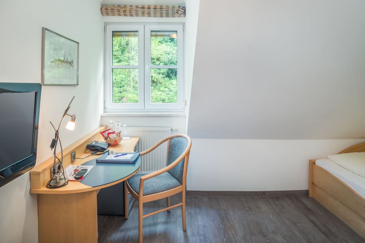 A small, tidy room with a wooden desk, a chair, a bed, a window with a view of greenery, and a framed picture on the wall.