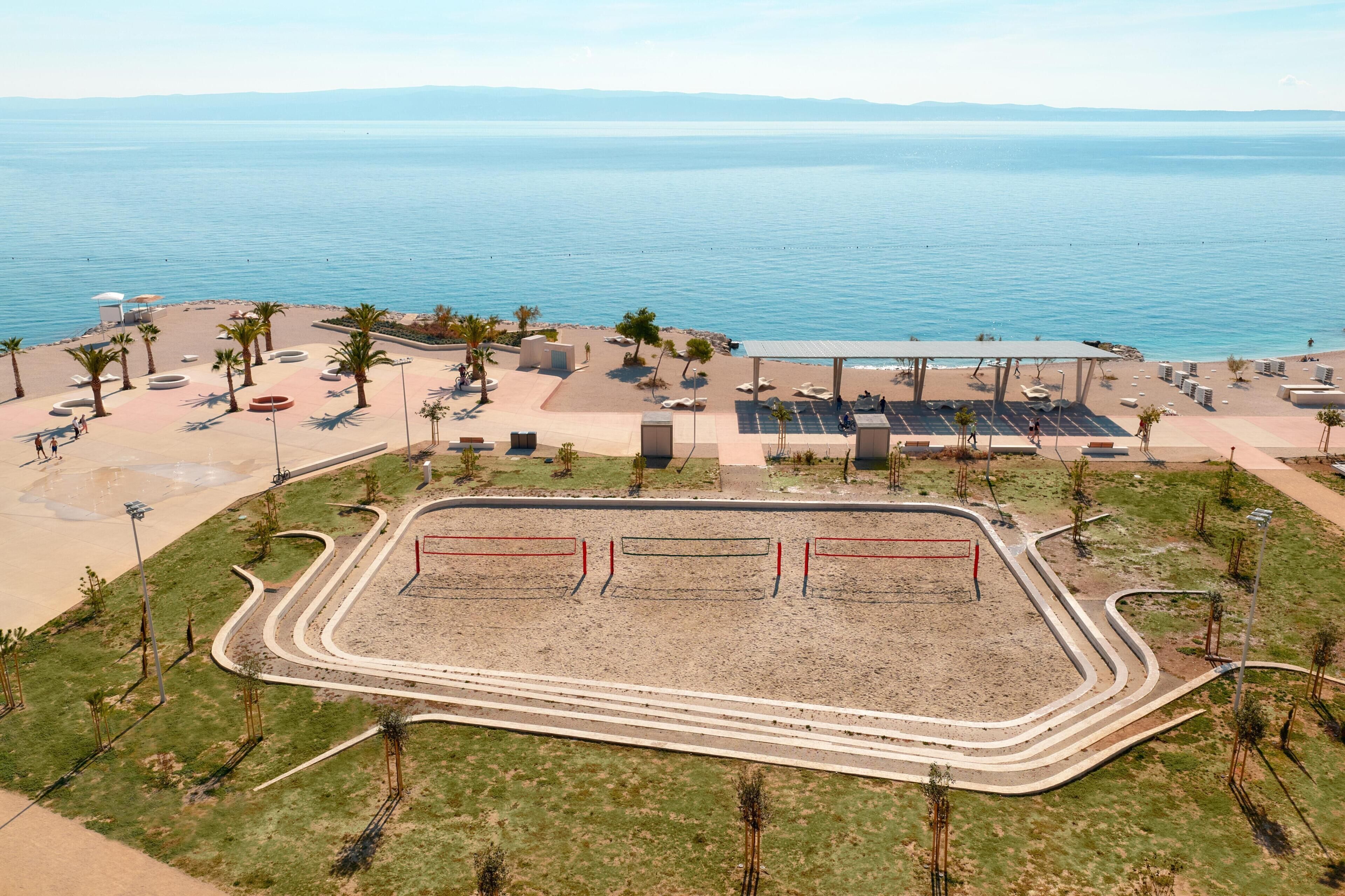 An aerial view of a beachside area with a sandy beach, palm trees, a seating area with umbrellas, and a fenced sports court.