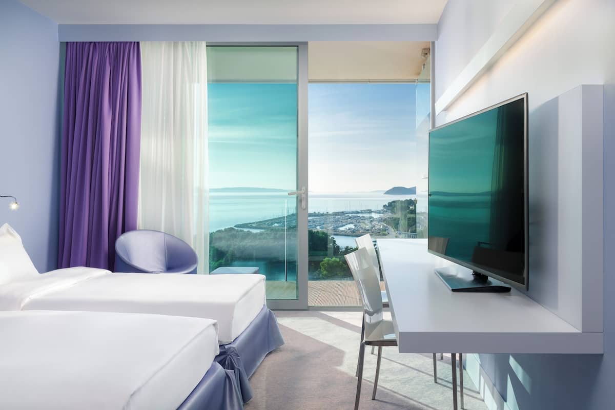 A modern hotel room with a large TV, a bed, a desk, and a view of the sea.