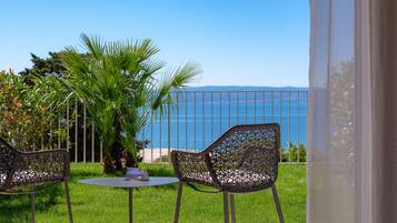 A balcony with two wicker chairs and a small table, overlooking a clear blue sea and a distant mountain range.