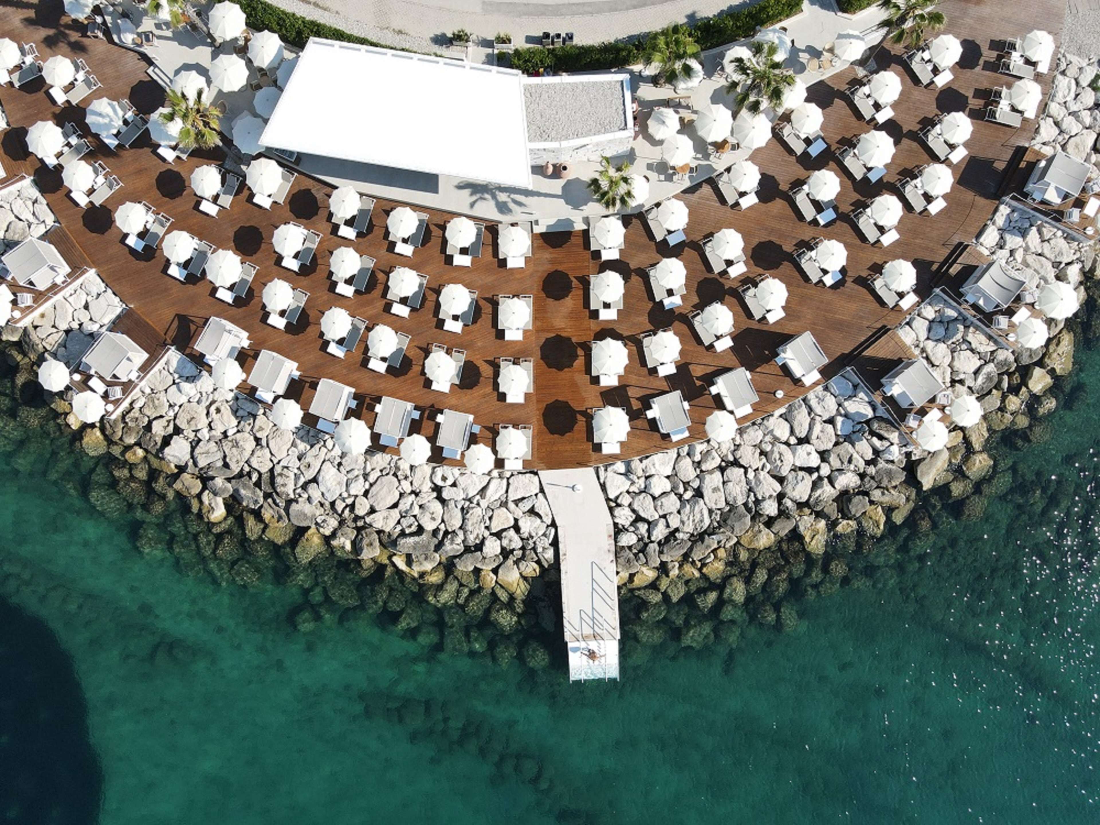 An aerial view of a resort with multiple sun loungers and umbrellas arranged around a central walkway, surrounded by a rocky shoreline.