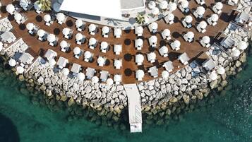An aerial view of a resort with multiple sun loungers and umbrellas arranged around a central walkway, surrounded by a rocky shoreline.