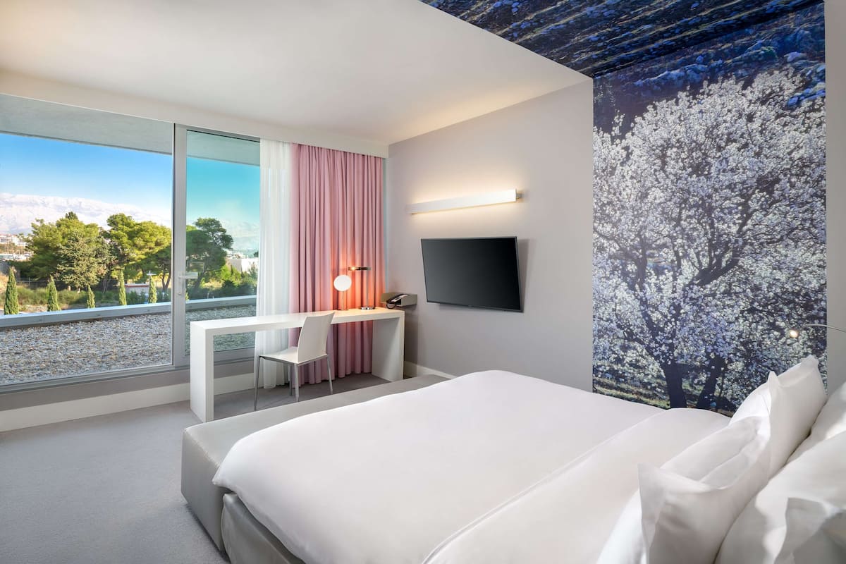 A modern hotel room with a large bed, a desk with a chair, a TV mounted on the wall, and a scenic view through the window.