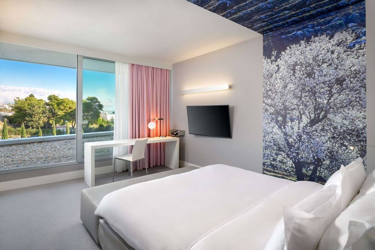A modern hotel room with a bed, desk, chair, and a TV on the wall.