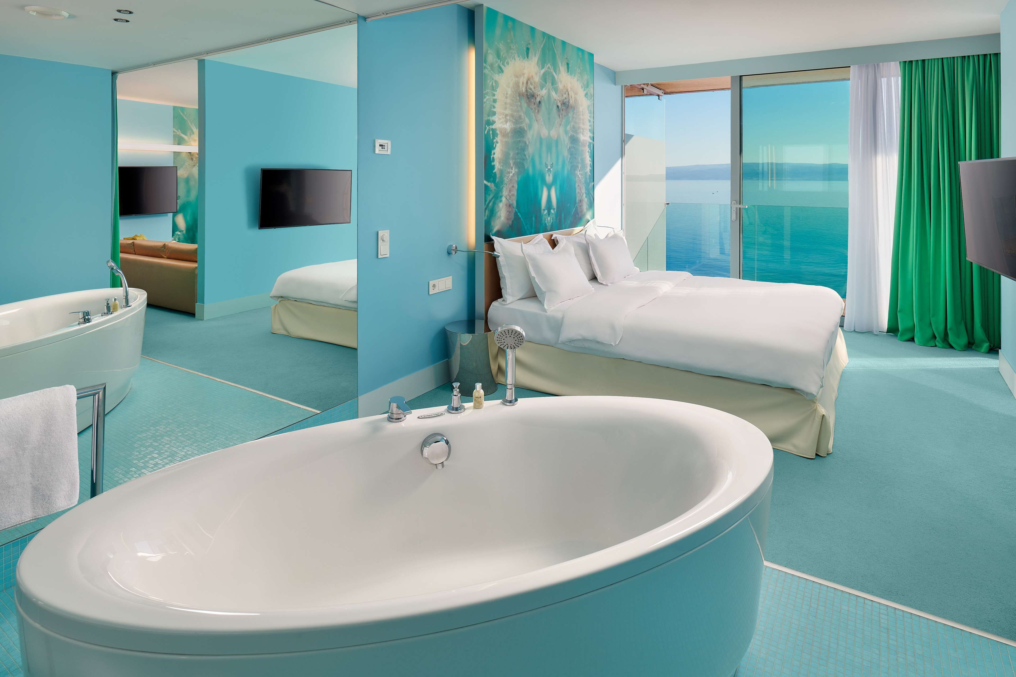 A modern hotel room with a large bathtub, a bed with white linens, a television, and a view of the sea.