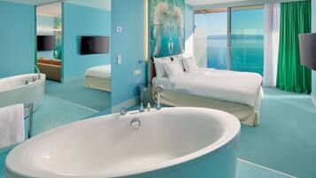 A modern hotel room with a large bathtub, a bed with white linens, a television, and a view of the sea.