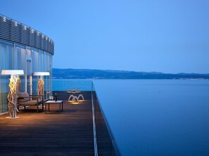 A modern outdoor deck with a glass balustrade overlooking a calm sea, featuring a seating area with a table, two lamps, and a wine glass.