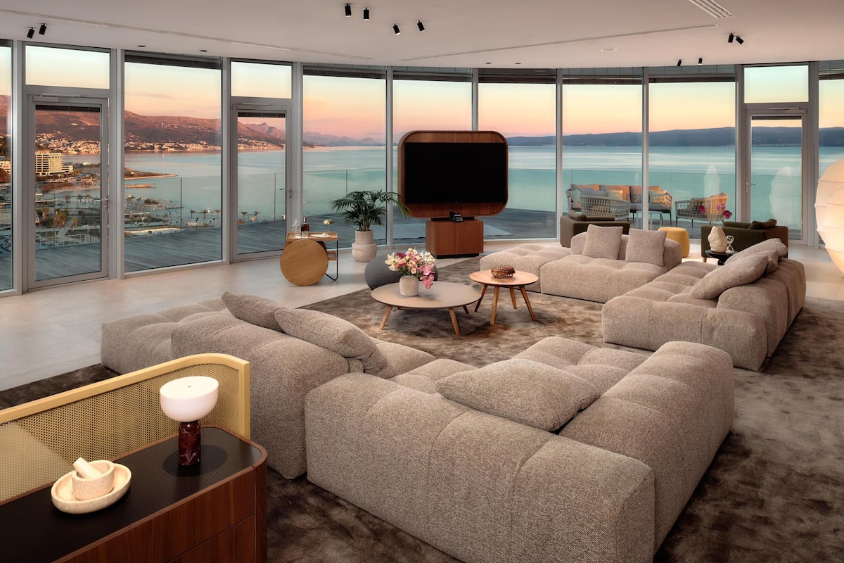 A modern living room with a large flat-screen TV, comfortable seating, a coffee table, and a view of a body of water.