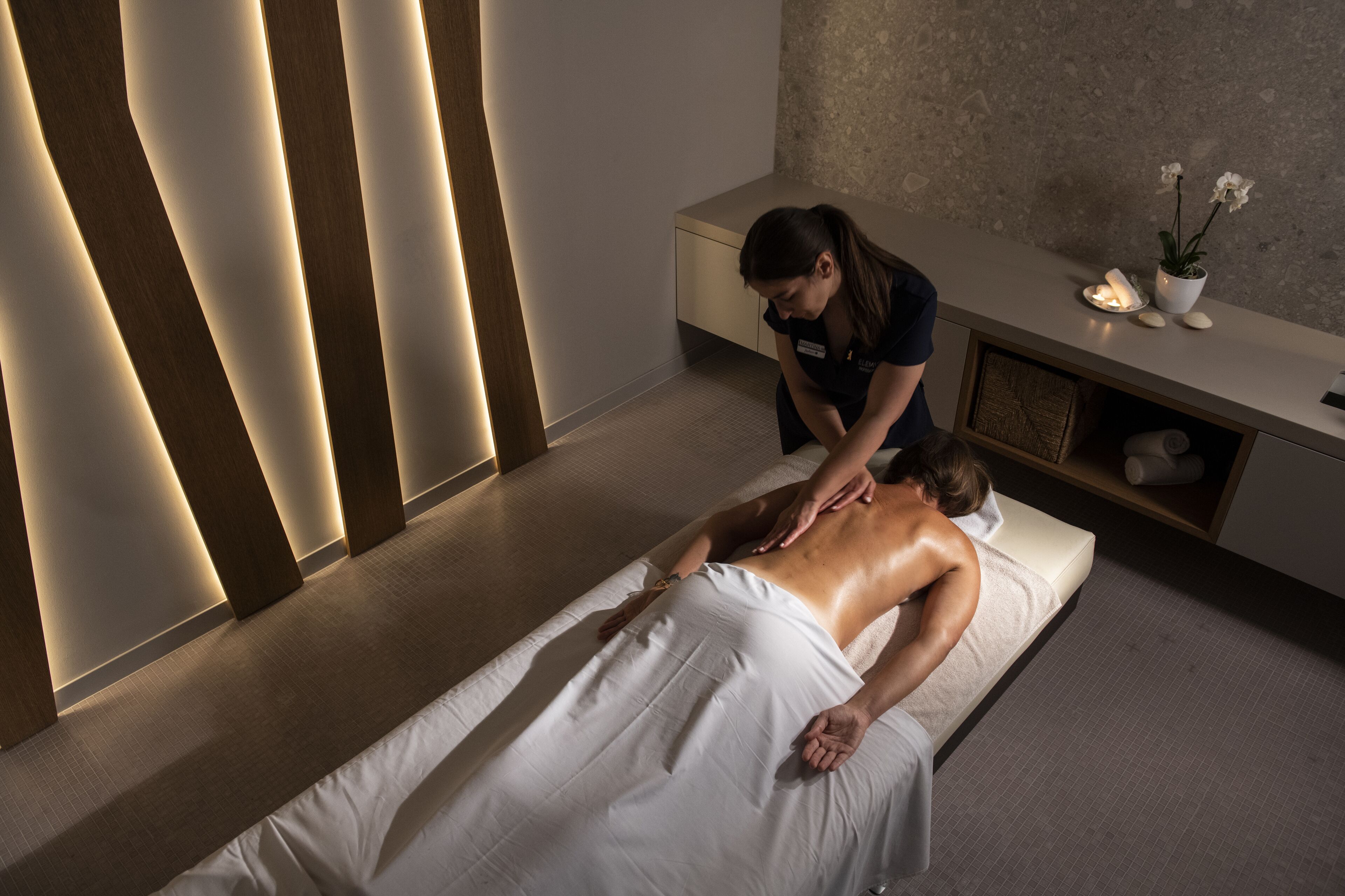 A massage therapy room with a massage table, a towel-covered client, a massage therapist, a small potted plant, and a wooden panel wall.