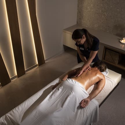 A massage therapy room with a massage table, a towel-covered client, a massage therapist, a small potted plant, and a wooden panel wall.