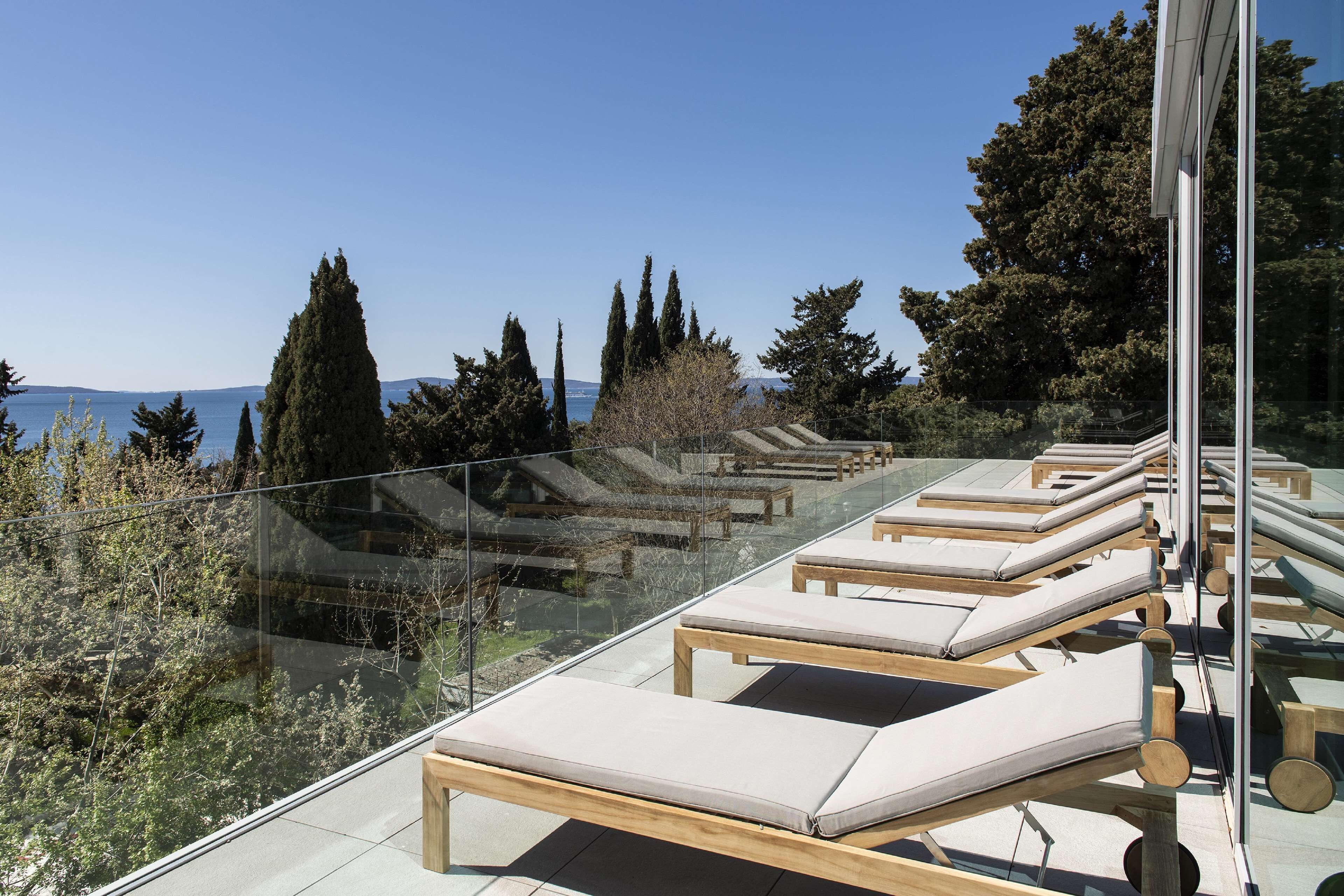 Una terraza en la azotea con tumbonas, barandillas de cristal y vistas al mar y a los árboles.