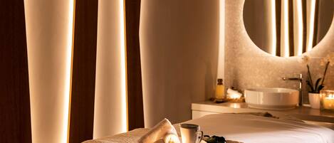 Une chambre d’hôtel avec un lit, une table de chevet sur laquelle se trouvent une tasse et une soucoupe, un miroir rond et une salle de bain équipée d’un lavabo et de bougies.