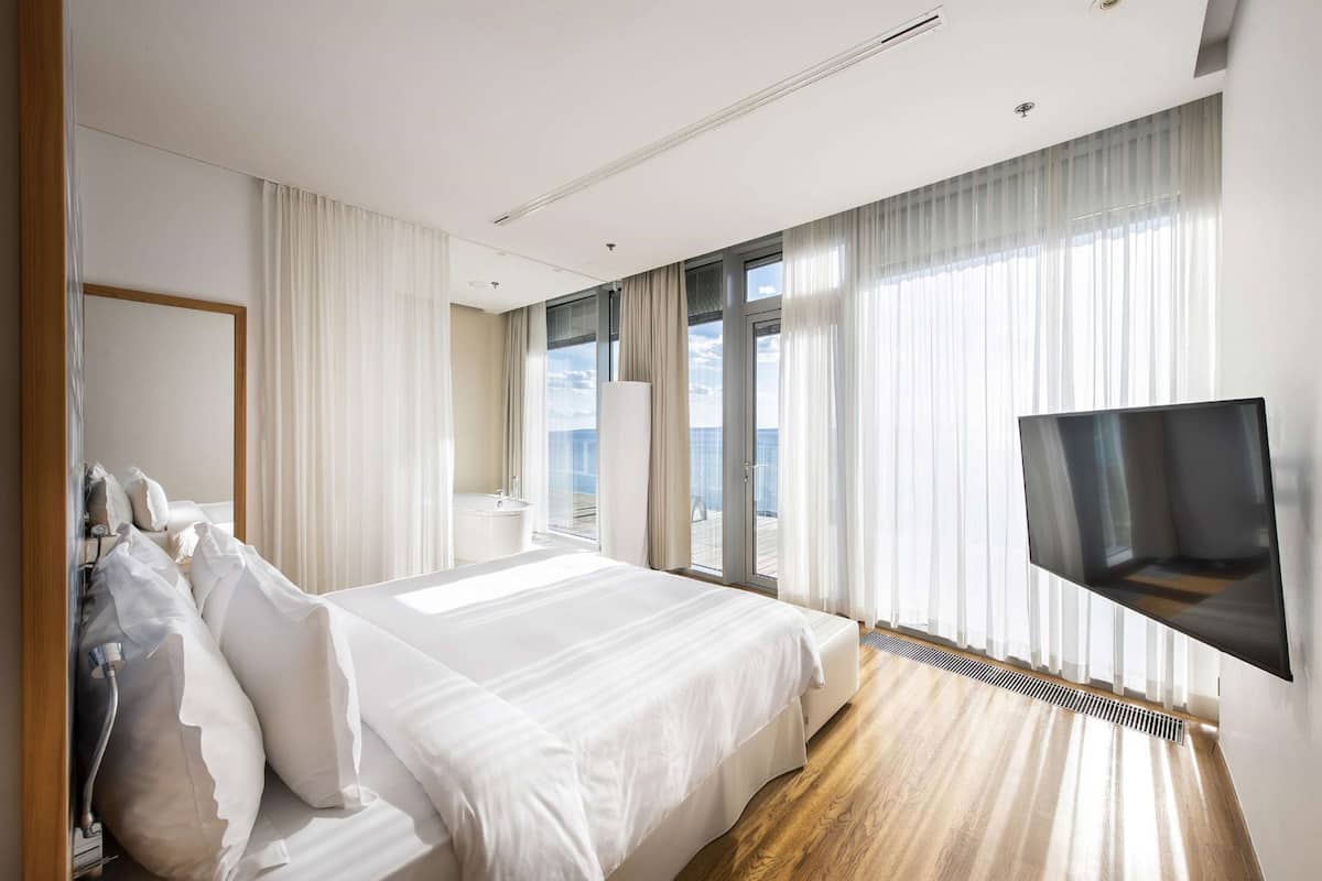 A modern hotel room with a large bed, a TV mounted on the wall, and a view of the sea through the windows.