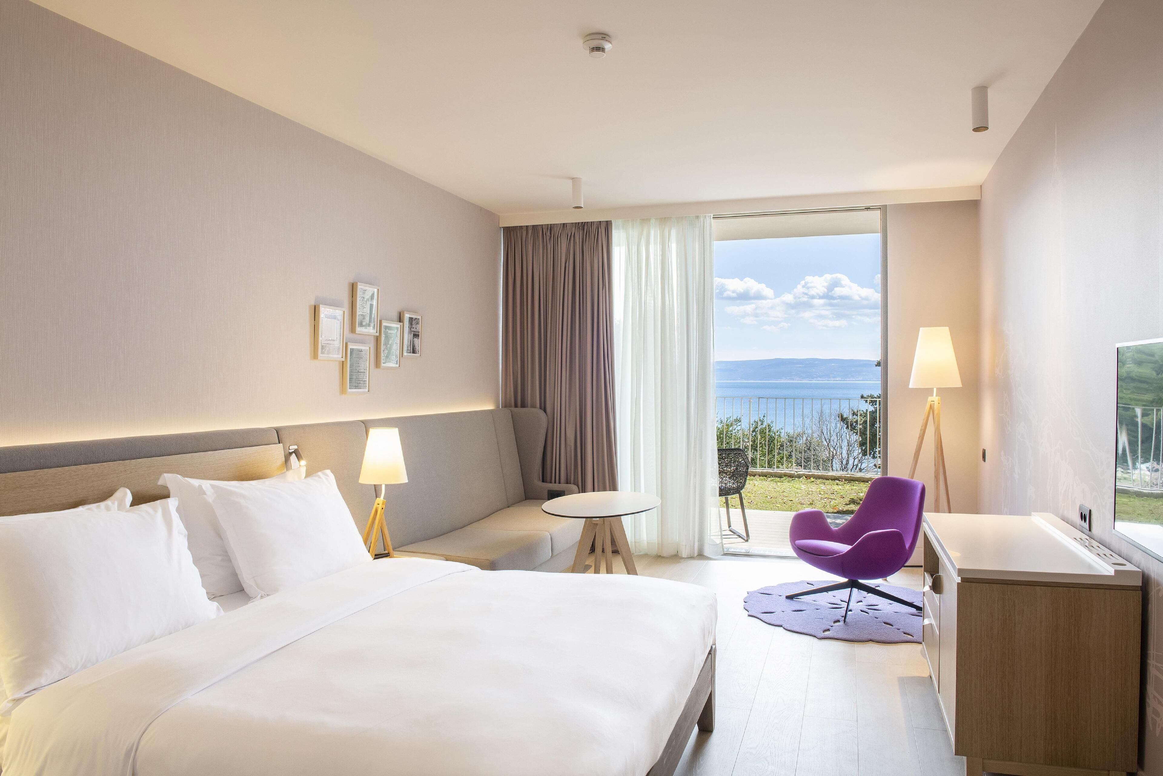A hotel room with a bed, a small table, a purple chair, a desk, and a view of the sea.
