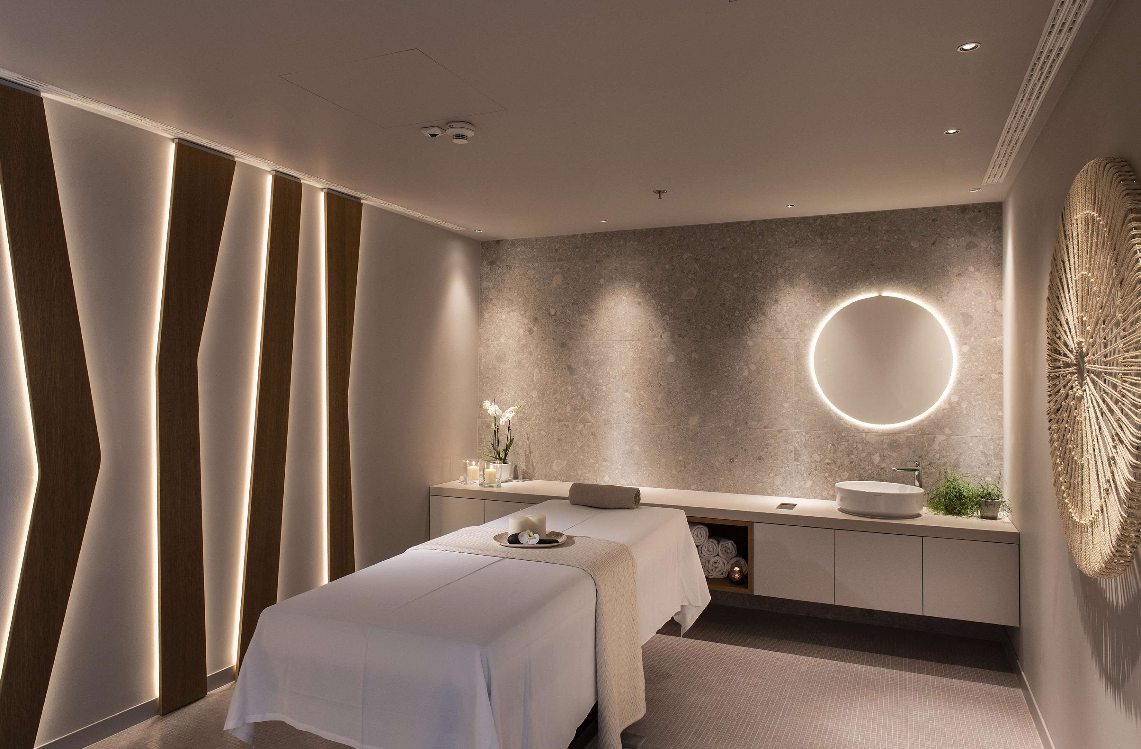 A modern spa room with a massage table, a round mirror, and a wall-mounted light fixture.