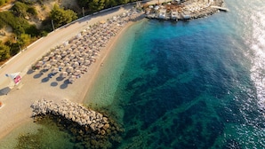 An aerial view of a beach with clear turquoise waters, a row of umbrellas, and a rocky breakwater.