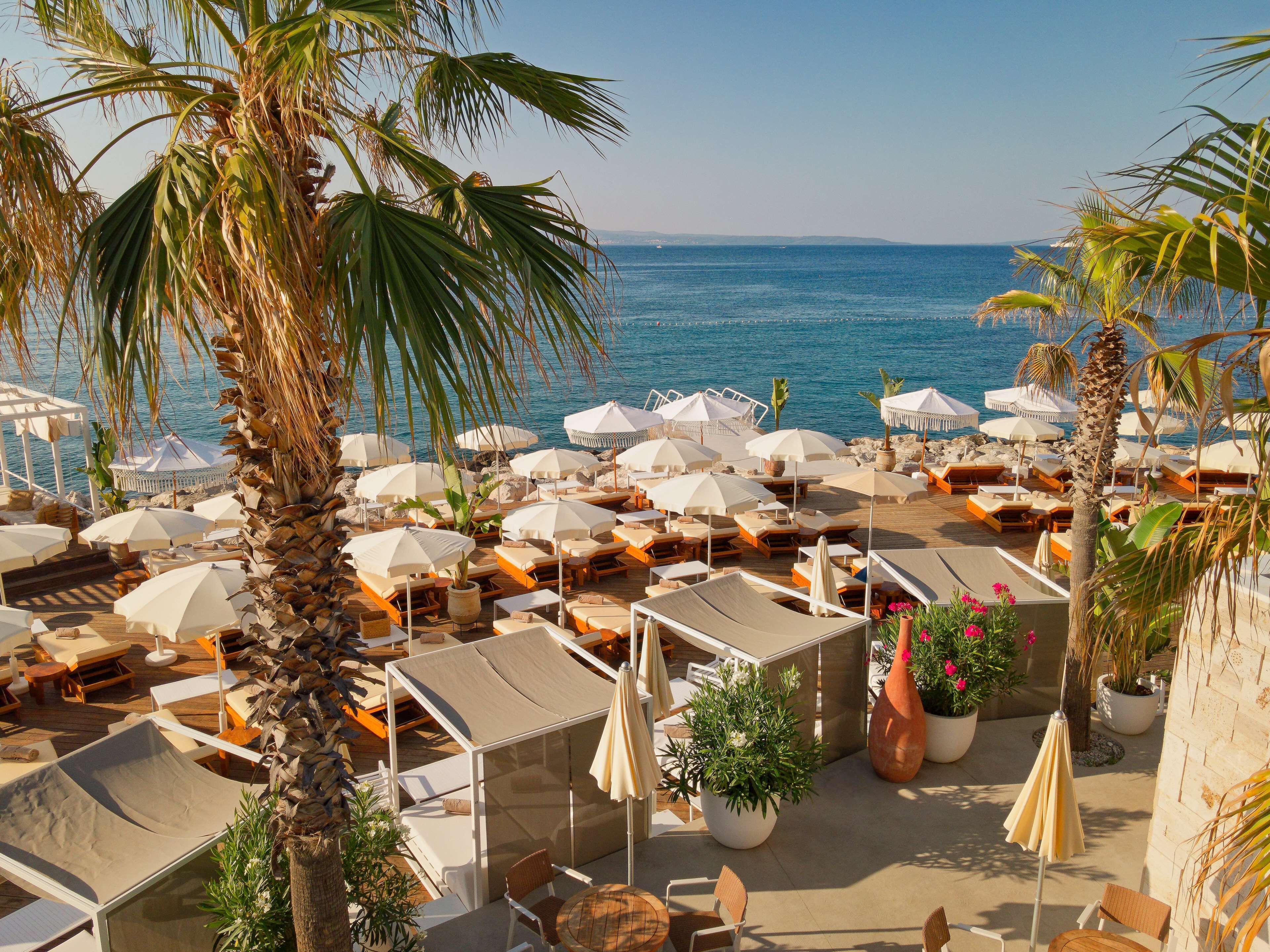 A beachside resort with multiple sun loungers and umbrellas, a pool area, and a view of the sea.