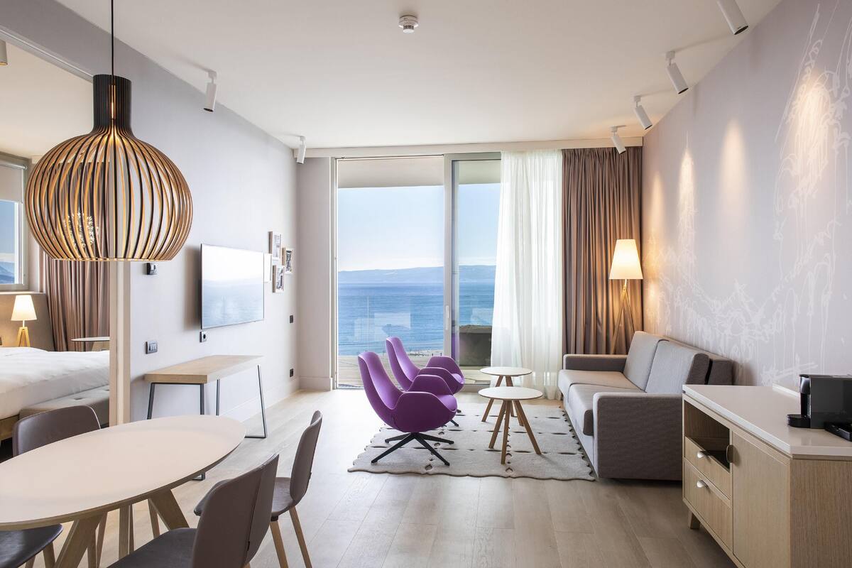 A modern hotel room with a large bed, a desk, a dining table, purple chairs, a sofa, and a view of the ocean.