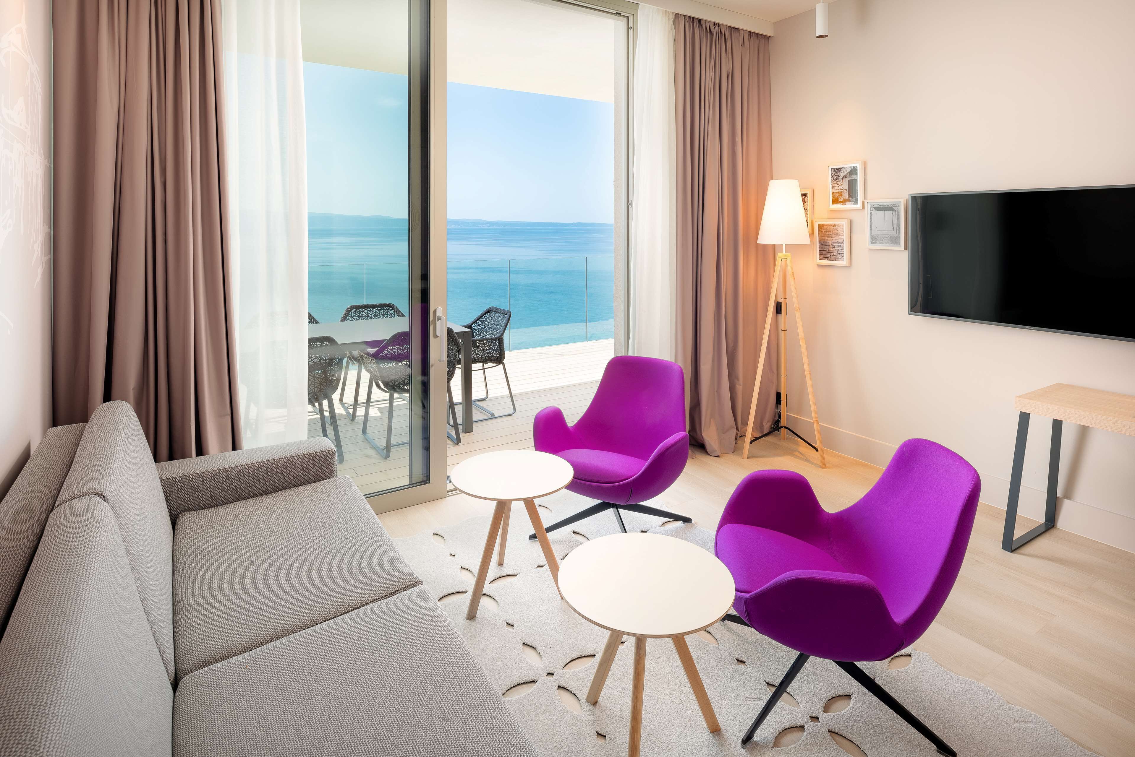 Een moderne hotelkamer met balkon, een bank, twee paarse stoelen, een klein tafeltje, een lamp en een televisie.