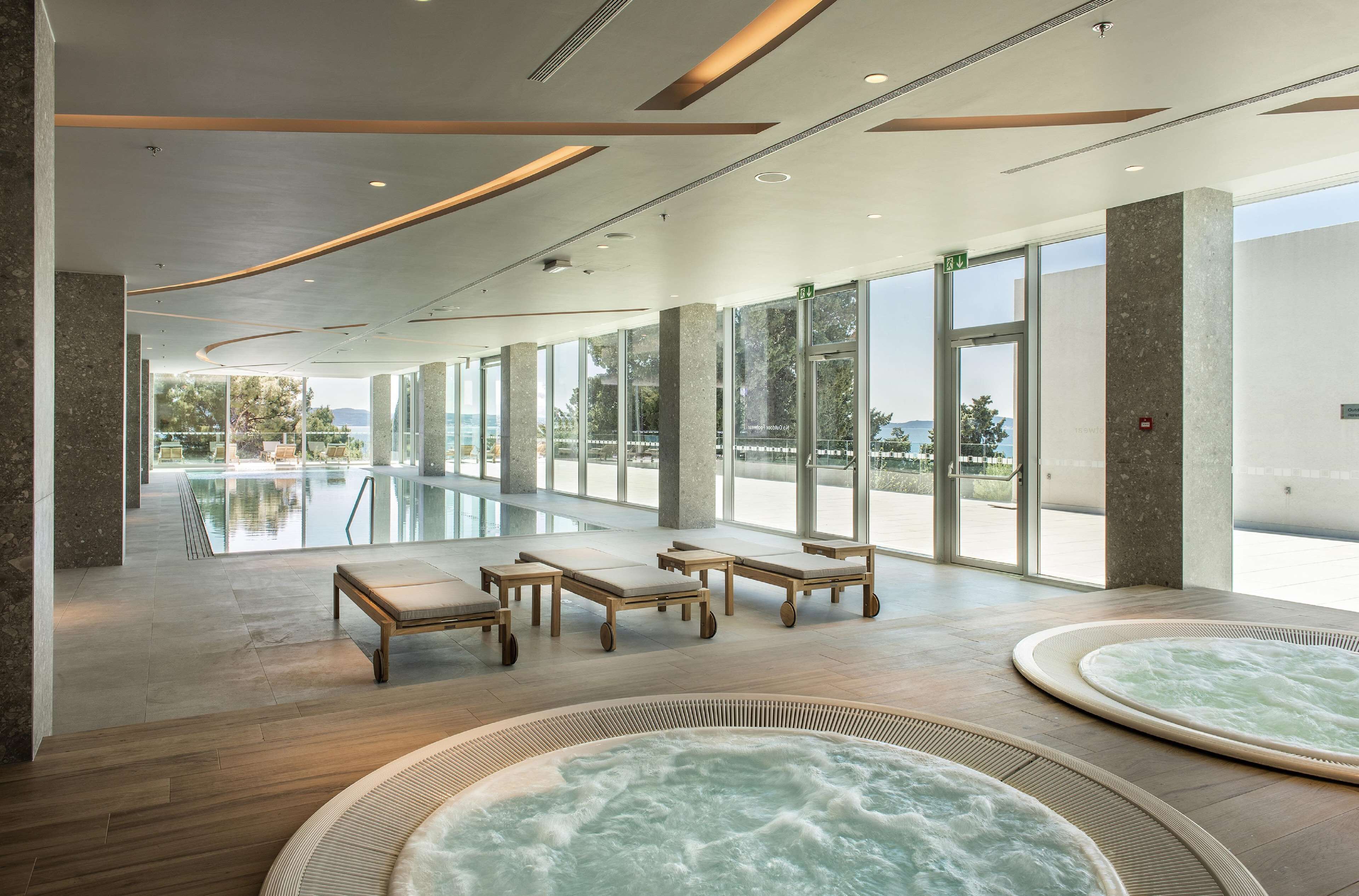 A spacious indoor pool area with lounge chairs and hot tubs, large windows, and a view of the outdoors.