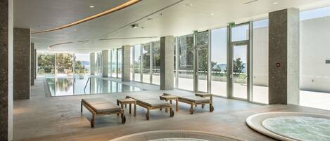 A spacious indoor pool area with lounge chairs and hot tubs, large windows, and a view of the outdoors.