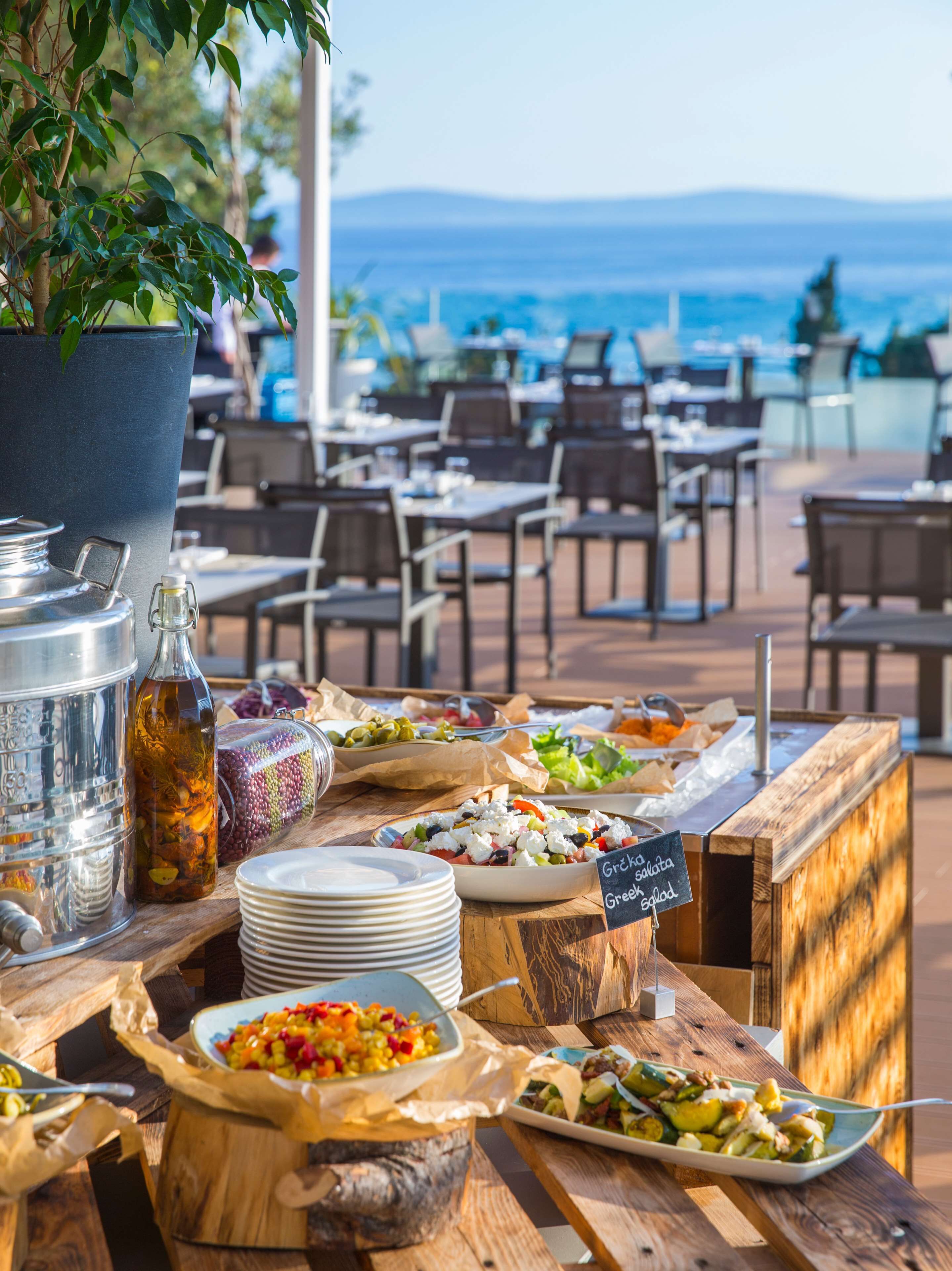 An outdoor dining area with various food dishes, a large metal container, and a bottle of drink. Tables and chairs are arranged facing a scenic view of the sea.