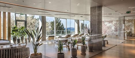 Un vestíbulo moderno con ventanas amplias, zonas de estar y plantas en macetas.