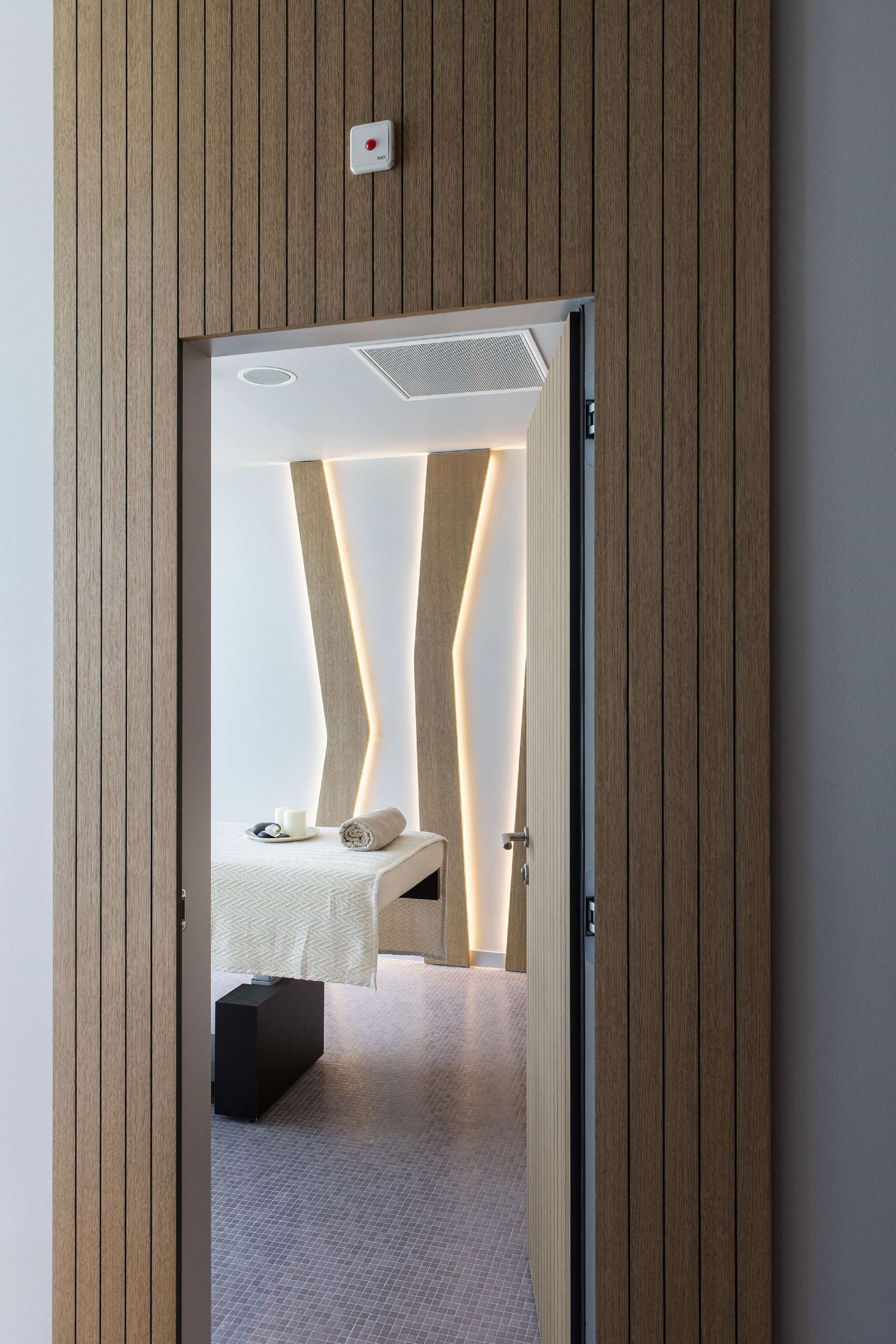 A modern hotel room with a wooden panel wall, a bed with a white headboard, a bedside table, and a small nightstand.