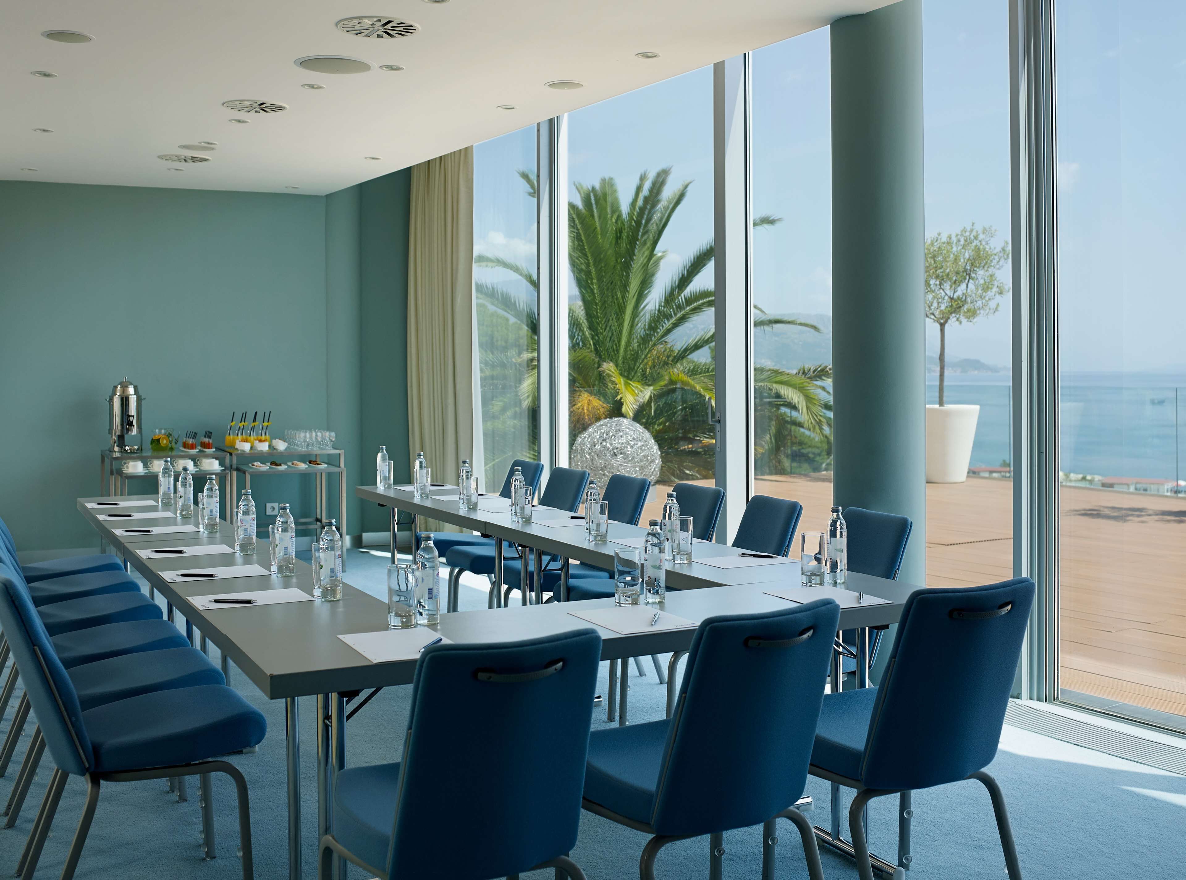 Uma sala de conferências com uma mesa comprida, cadeiras azuis e vista para a praia e palmeiras através de grandes janelas.