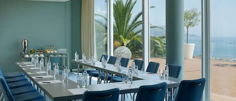 Uma sala de conferências com uma mesa comprida, cadeiras azuis e vista para a praia e palmeiras através de grandes janelas.