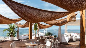 An outdoor seating area with tables and chairs, a wooden deck, and a view of the sea.