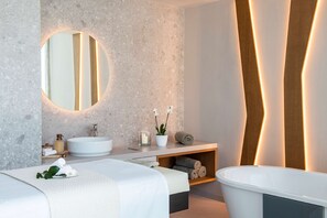 A modern bathroom with a round mirror, two sinks, a bathtub, and a wooden shelf with towels.