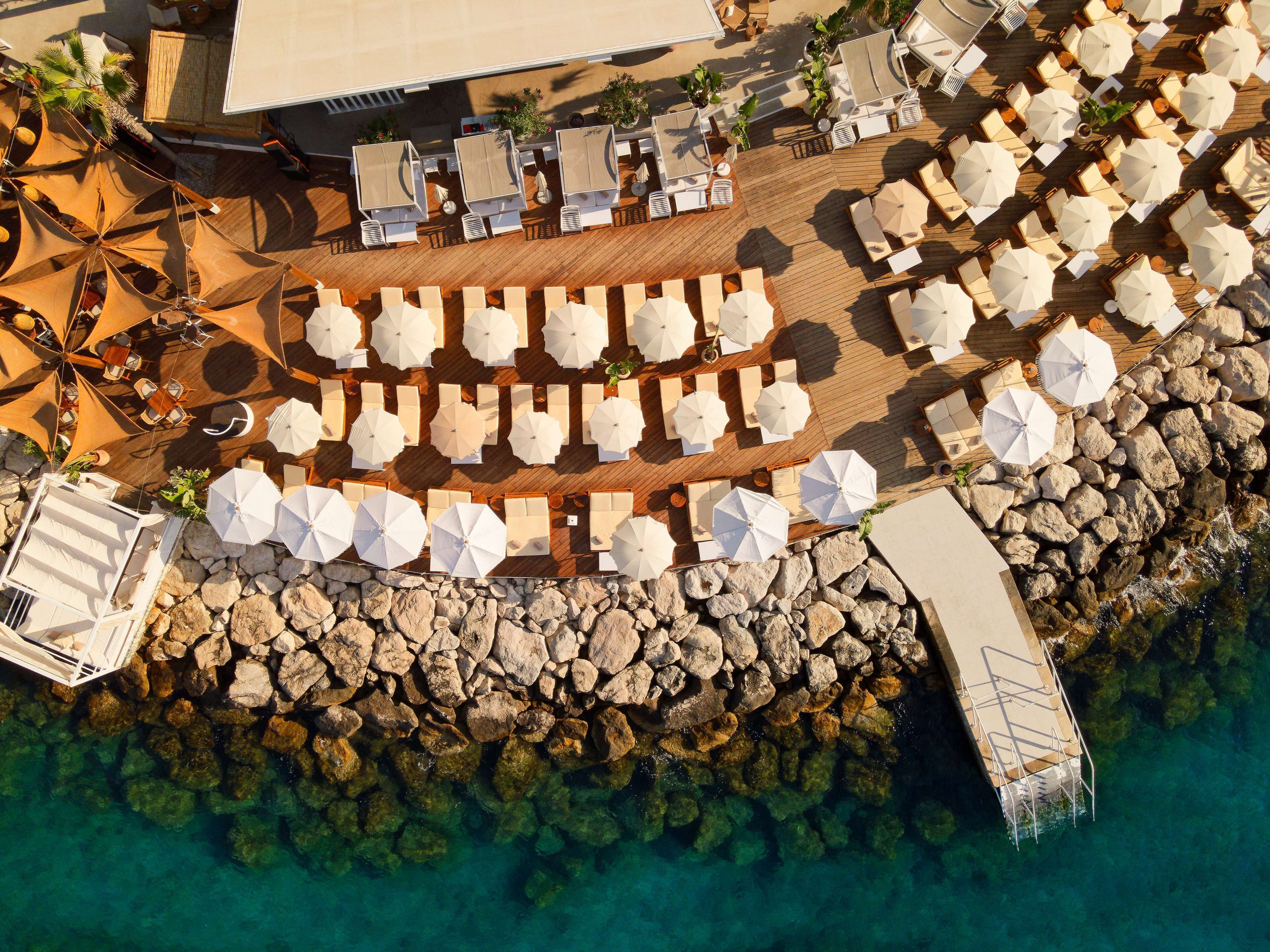 An aerial view of a beachfront resort with multiple sun loungers and umbrellas, a wooden deck, and a rocky shoreline.