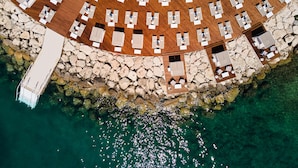 An aerial view of a coastal hotel with multiple sun loungers arranged in rows, a rocky shoreline, and clear turquoise water.