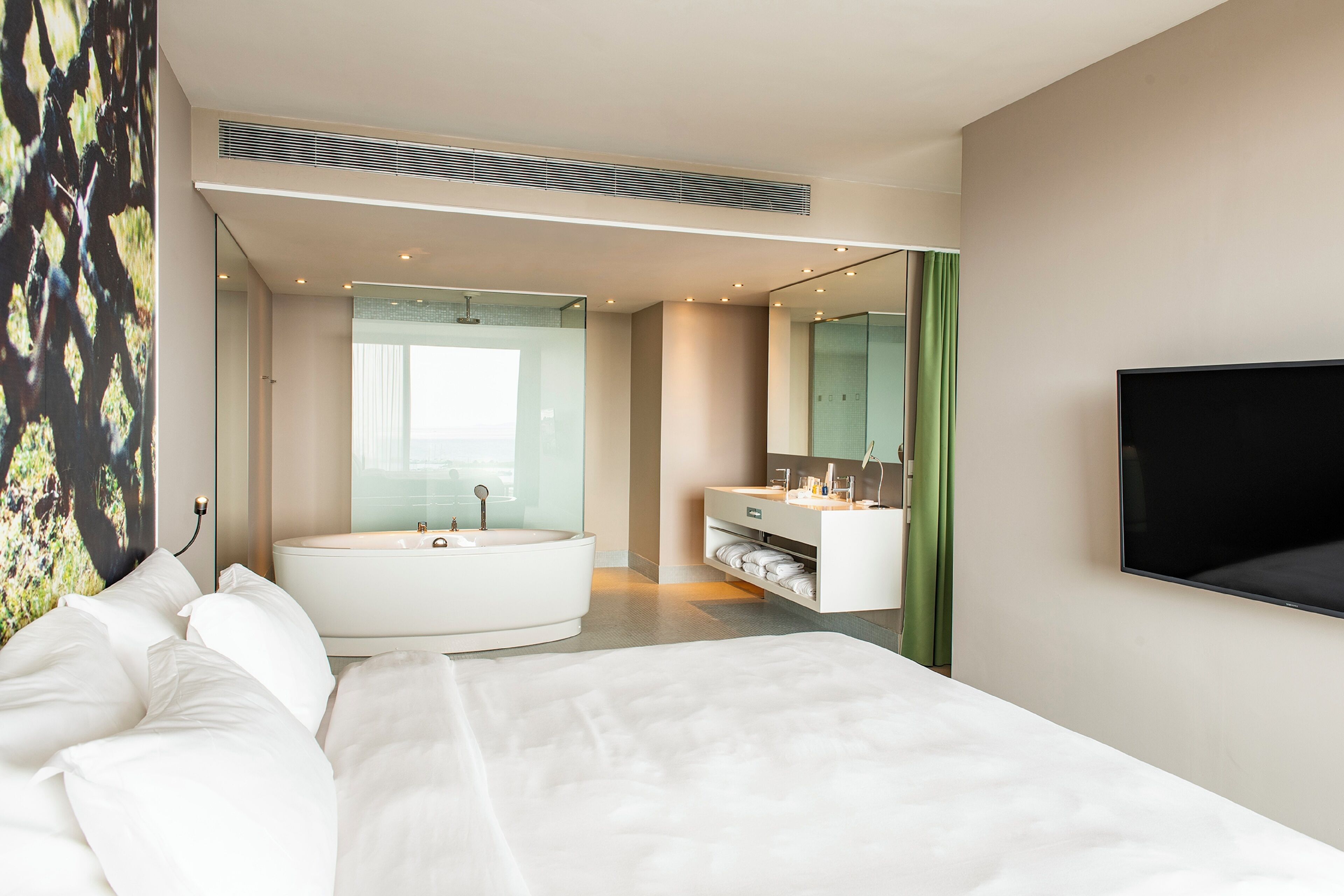 A modern hotel room with a large bed, a separate bathroom with a bathtub, a flat-screen TV, and a large window.