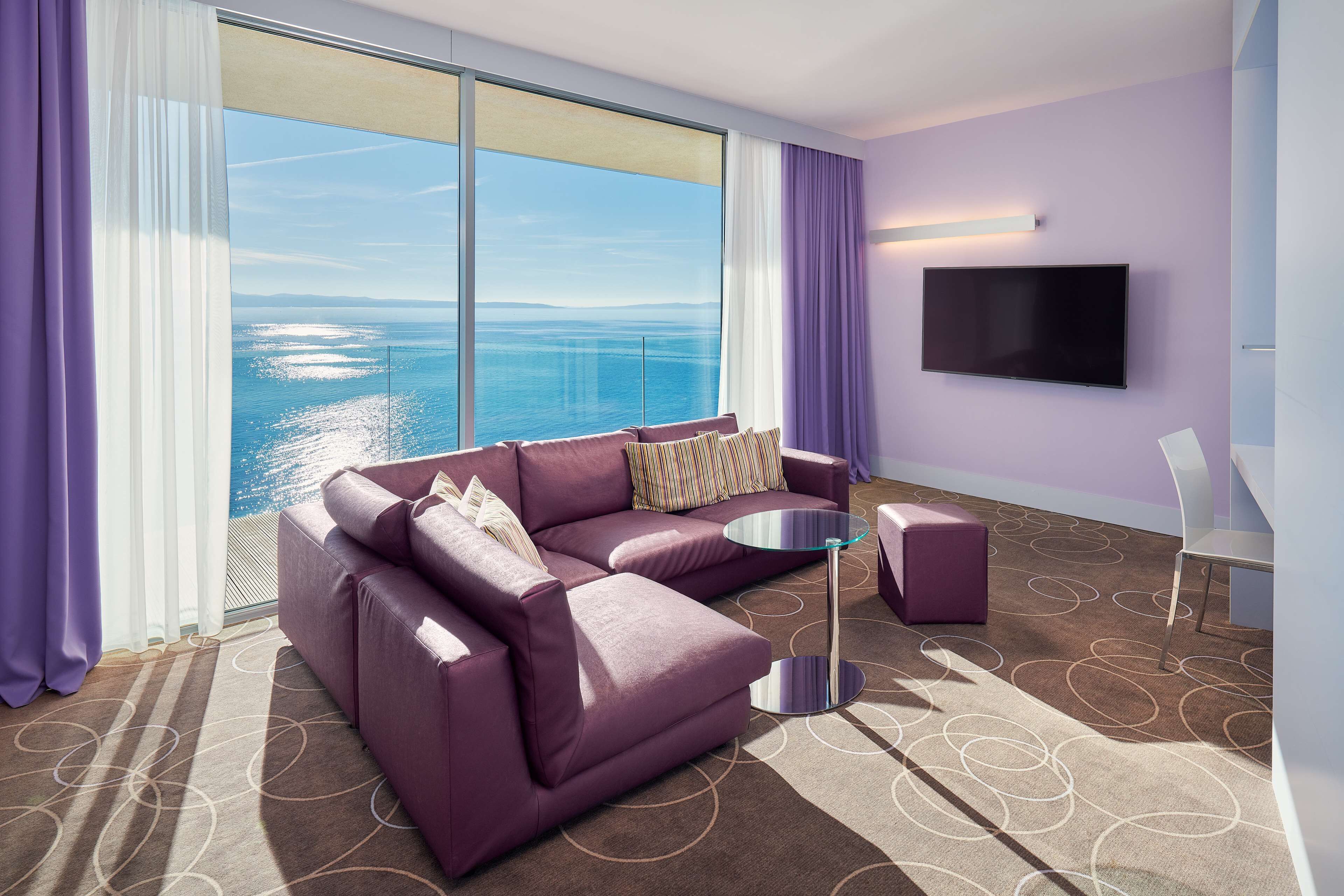 A modern hotel room with a large window offering a view of the sea, a purple sofa, a small round table, and a flat-screen TV mounted on the wall.