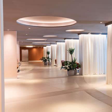 A modern hotel lobby with a long corridor, large white curtains, and potted plants.