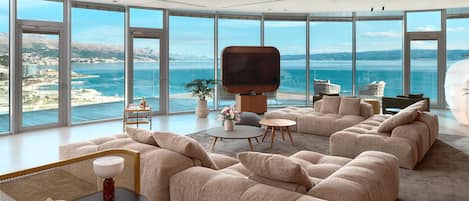 A modern living room with a large TV, comfortable seating, and a view of the sea.