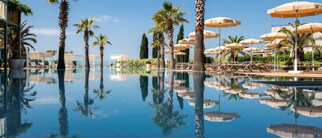Zona de piscina con palmeras y sombrillas, rodeada de sillones de descanso y un cielo despejado.