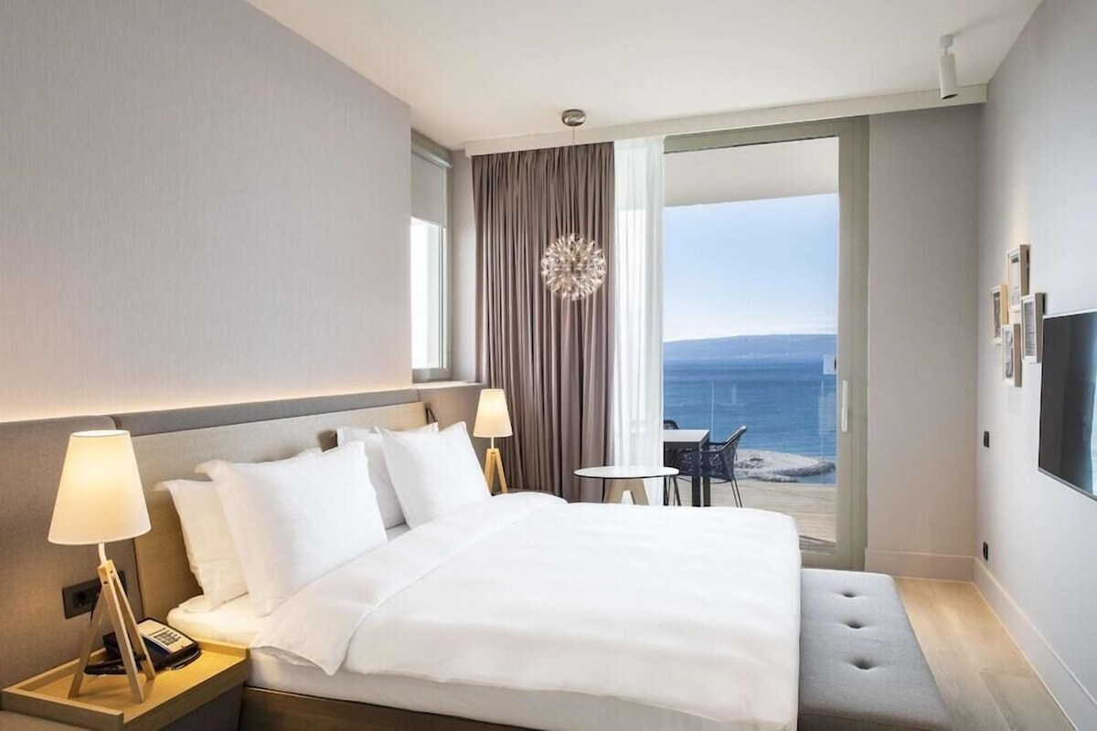 A hotel room with a bed, bedside lamps, a TV, a balcony with a view of the sea, and a small table.