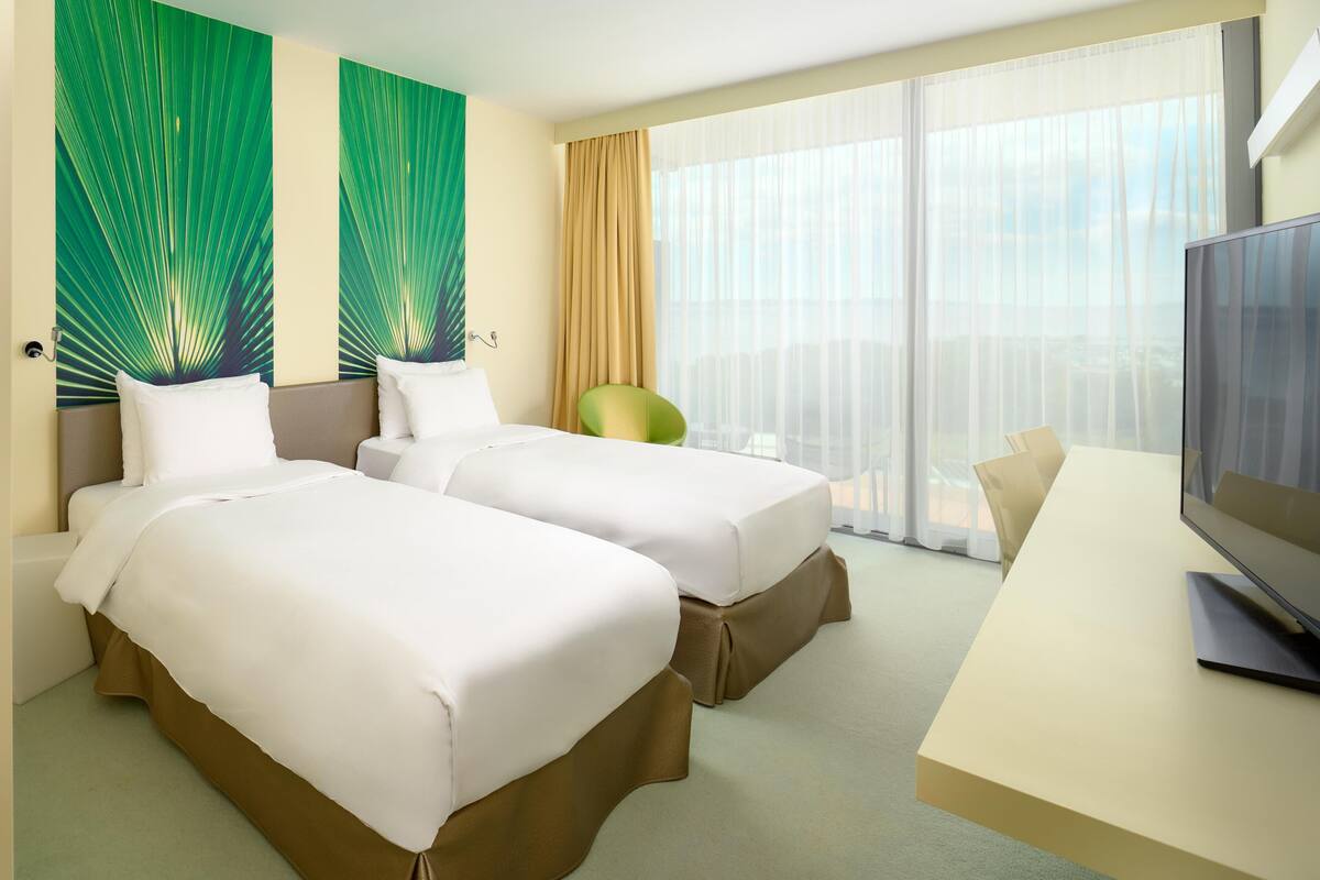 A hotel room with two beds, a desk, a TV, and a large window with curtains.