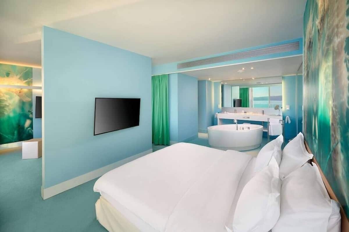 A modern hotel room with a large bed, a flat-screen TV, a bathtub, and a bathroom with a large mirror.