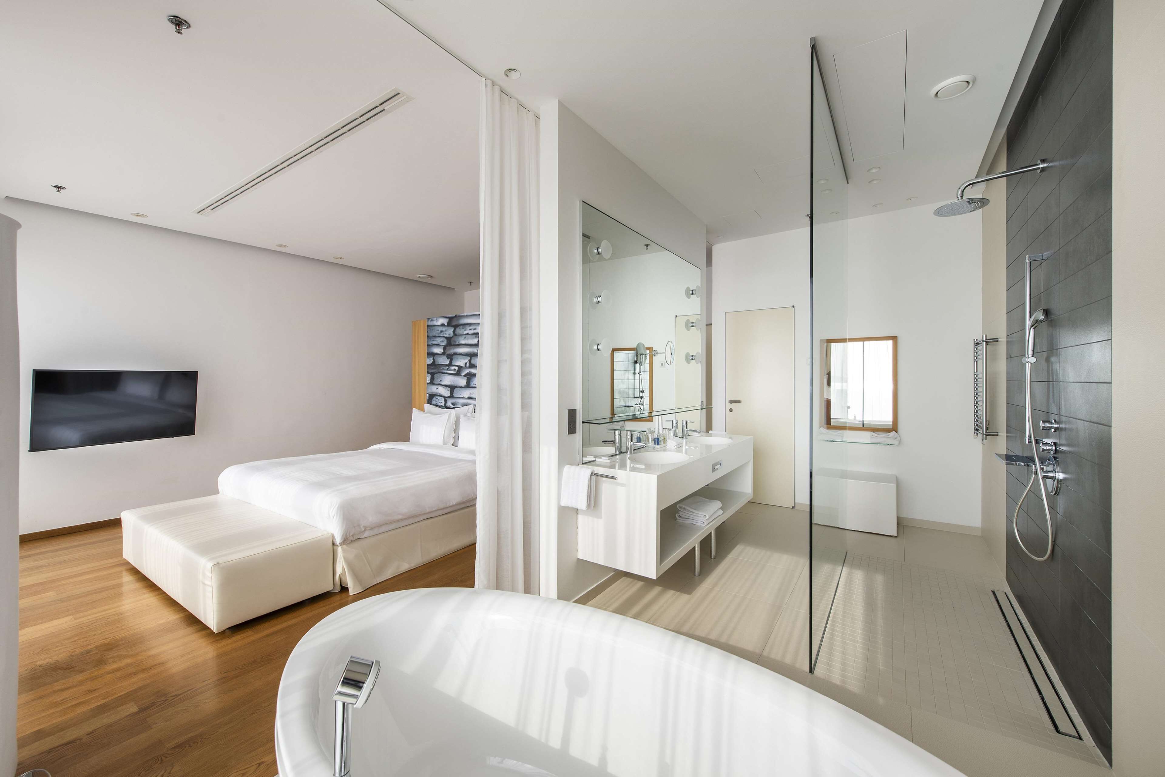A modern hotel room with a large bed, a separate seating area, a bathroom with a shower, and a flat-screen TV mounted on the wall.